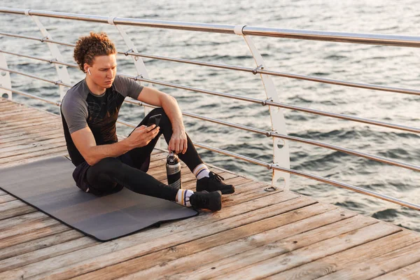 Portrait of a calm sportsman sitting on a fitness mat the beach, listening to music with earphones, holding mobile phone