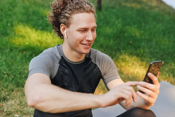 微笑的年轻运动员听音乐与耳机 而坐在户外健身垫 手持手机 — 图库照片