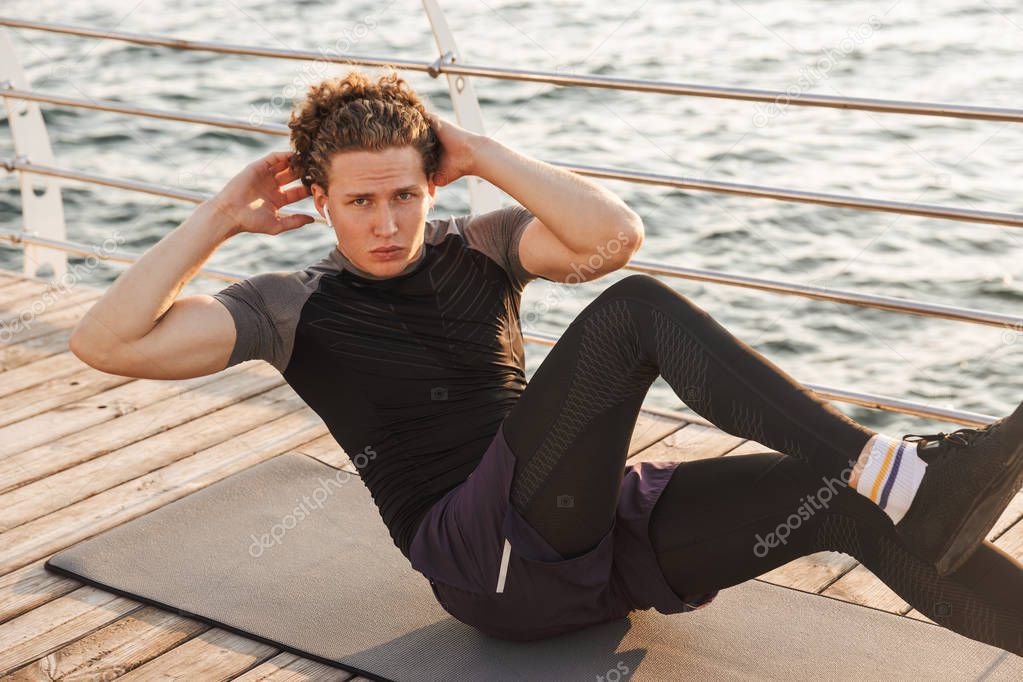 Portrait of a focused sportsman doing exercises on a fitness mat the beach, listening to music with earphones