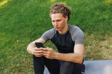 Cep telefonu tutan bir fitness mat açık havada, otururken kulaklık ile müzik dinlemek çekici genç sporcu