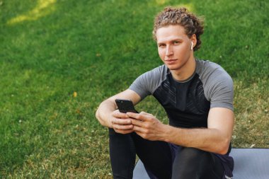 Genç sporcu bir fitness mat açık havada, otururken kulaklık ile müzik dinlerken cep telefonu tutan gülümseyerek