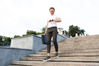 Mutlu kıvırcık iş adamı merdivenlerde yürürken kol saati görünümlü evrak çantası ile tam boy görüntü