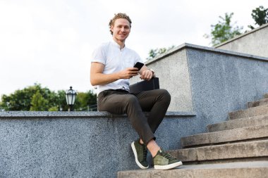 Kıvırcık iş adamı evrak çantası holding smartphone ile gülümseyen ve merdivenlerden yakınındaki otururken kameraya bakıyor