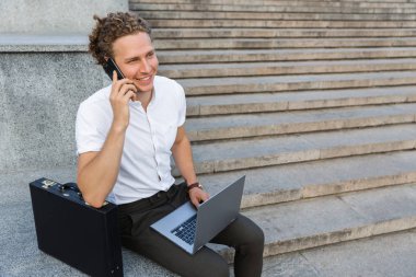 Gülümseyen kıvırcık iş adamı merdivenlerden yakınındaki otururken smartphone ile konuşurken evrak çantası ve dizüstü bilgisayar ile