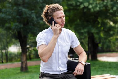 Ciddi kıvırcık iş adamı ile smartphone ile konuşmak ve uzak parkta bankta otururken seyir evrak çantası