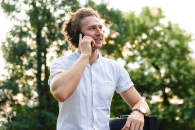 Memnun kıvırcık iş adamı ile smartphone ile konuşmak ve uzak parkta bankta otururken seyir evrak çantası