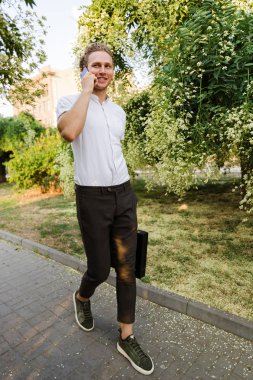 Gülen kıvırcık iş adamı sokakta yürürken smartphone ile konuşurken evrak çantası ile tam boy görüntü