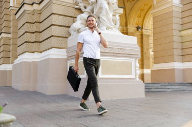 Mutlu kıvırcık iş adamı sokakta yürürken smartphone ile konuşurken evrak çantası ile tam boy görüntü