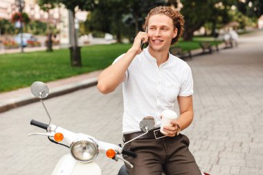 Smartphone ile konuşmak ve uzakta scooter açık havada üzerinde otururken bakarak mutlu kıvırcık iş adamı