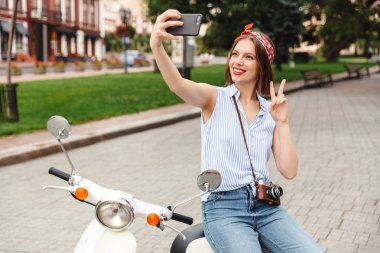Selfie smartphone yapma ve açık havada scooter üzerinde otururken barış hareketi gösterilen mutlu genç hipster kadın