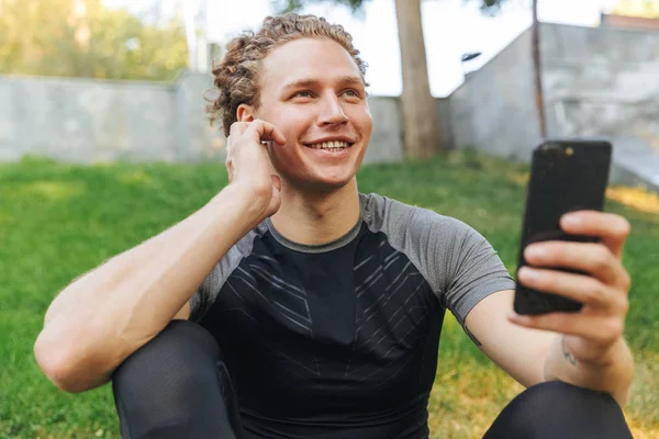 快乐的年轻运动员听音乐与耳机 而坐在户外健身垫 手持手机 — 图库照片