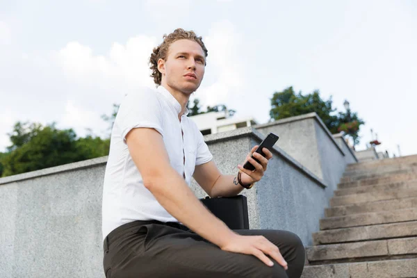 Ernster Lockiger Geschäftsmann Mit Aktentasche Smartphone Der Hand Und Wegschauend — Stockfoto