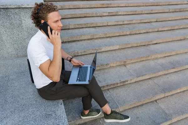 穏やかな巻き毛ビジネスの男性のスマート フォンでは話しながら階段のそばに座ってブリーフケース ラップトップ コンピューターとの側面図 — ストック写真