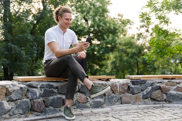 Mutlu kıvırcık iş adamı ile parkta bankta otururken smartphone kullanarak evrak çantası