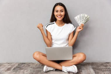 Image of excited happy young woman sitting isolated over grey background using laptop computer holding money make winner gesture.