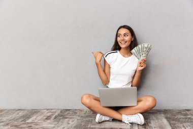 Image of excited happy young woman sitting isolated over grey background using laptop computer holding money looking aside pointing.