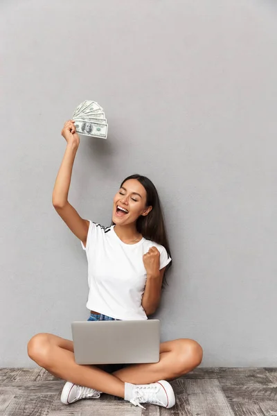 分離ラップトップ コンピューターお金を使用して灰色の背景の上に座って興奮して幸せな若い女の画像作る勝者ジェスチャー — ストック写真