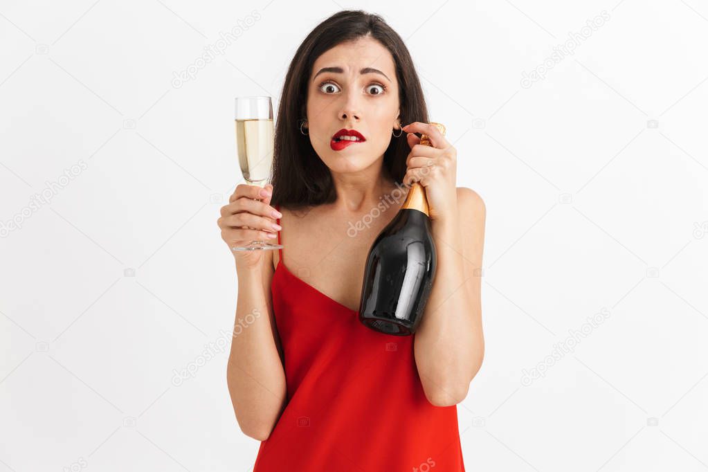Portrait of a frustrated young woman in dress holding glass and bottle of champagne isolated over white background