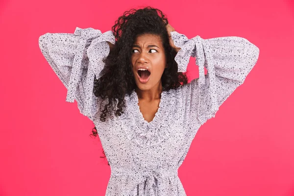 Portrait of a shocked young african woman in summer dress standing isolated over pink background, screaming, looking away