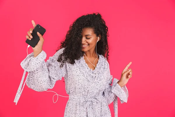 一个微笑的年轻非洲妇女的肖像在夏季礼服站在粉红色背景孤立 听音乐与耳机 手持手机 — 图库照片