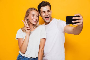 Fotoğraf mutlu seven Çift sarı duvar arka plan üzerinde izole duran genç bir selfie cep telefonuyla almak heyecanlı olun barış hareketi.