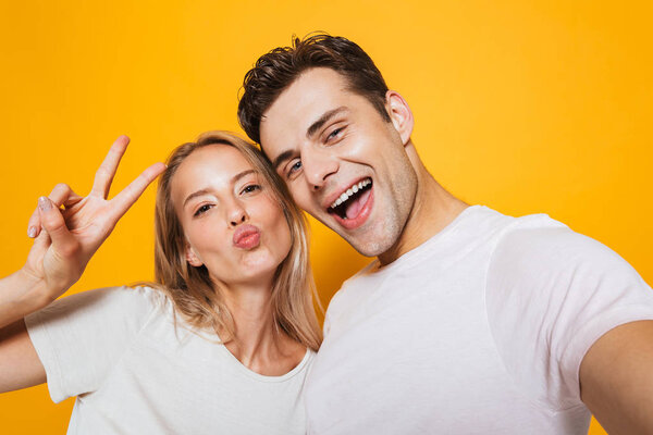 Photo of happy excited young loving couple standing isolated over yellow wall background take a selfie by camera showing peace gesture.