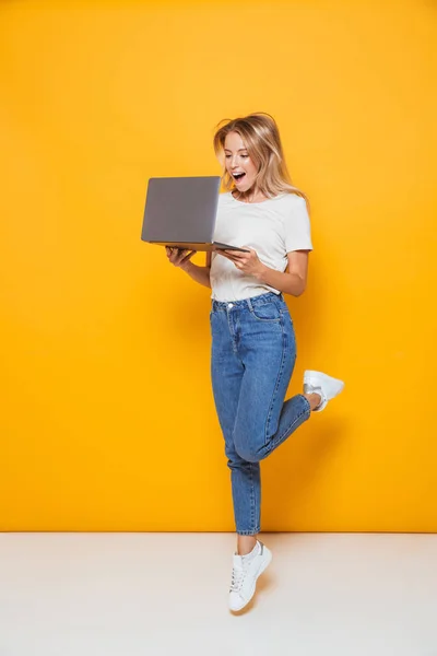 Photo of happy excited young woman jumping isolated over yellow wall background using laptop computer.