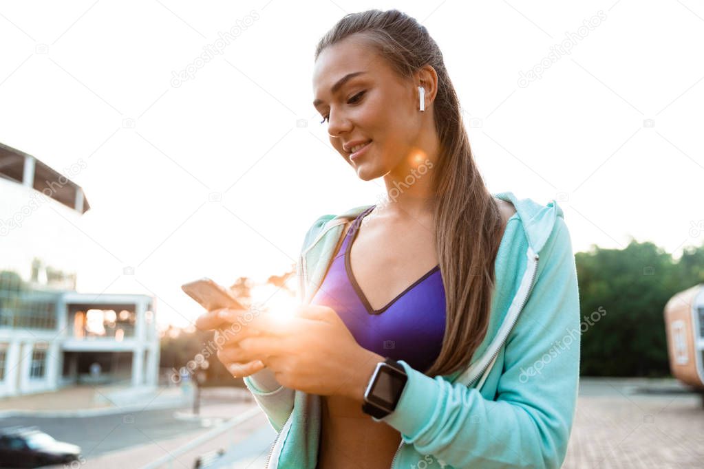 Photo of pretty young sports woman in park outdoors listening music with earphones using mobile phone.