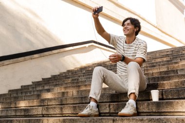 Yakışıklı genç adam açık havada müzik dinleme basamaklarında oturan görüntüsünü almak bir selfie cep telefonuyla.