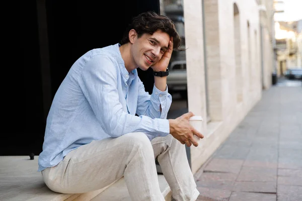 Uśmiechający Się Młody Człowiek Dorywczo Picia Kawy Siedząc Przy Ulicy — Zdjęcie stockowe