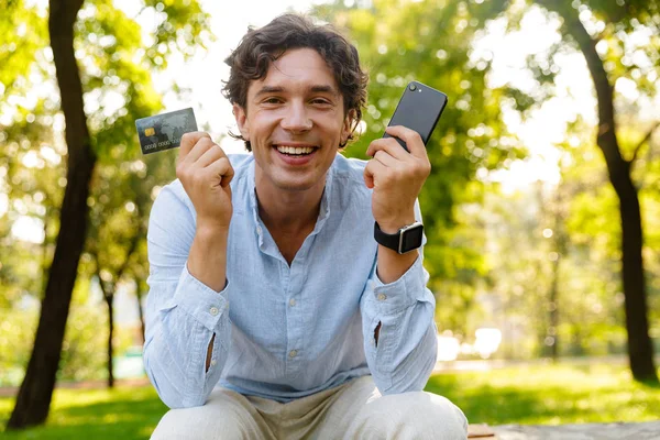 兴奋的年轻休闲男子手持手机 而坐在城市公园 显示信用卡 — 图库照片