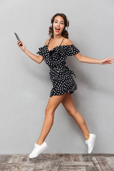 Cep telefonu tutan kulaklık ile müzik dinlemek neşeli bir genç kız elbise gri arka plan üzerinde atlama tam uzunlukta portresi