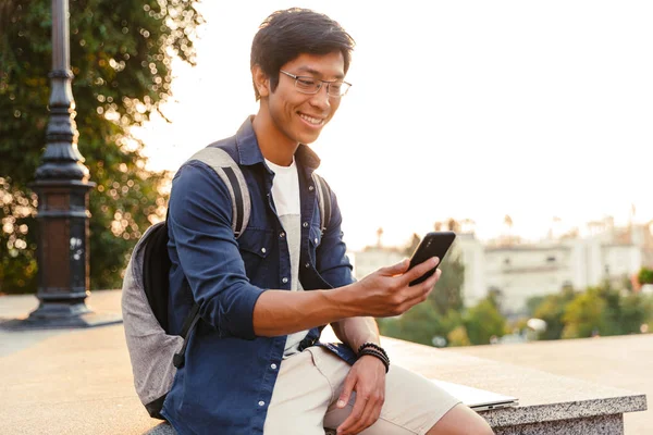 Joyful Asiático Estudante Masculino Óculos Usando Smartphone Enquanto Sentado Parque — Fotografia de Stock