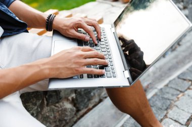 Asyalı erkek öğrenci parkta bankta otururken dizüstü bilgisayar kullanarak kırpılmış görüntü