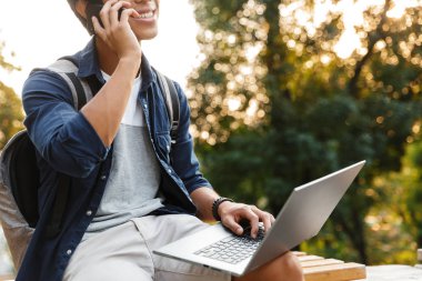 Gülen Asya erkek öğrenci dizüstü bilgisayar ile parkta otururken smartphone ile konuşurken kırpılmış görüntü