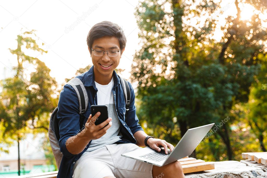 Smiling asian male student in eyeglasses using smartphone while sitting on bench in park with laptop computer