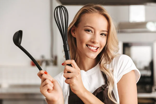 Smiling young woman chef cook in apron standing at the kitchen, holding ladle and whisk