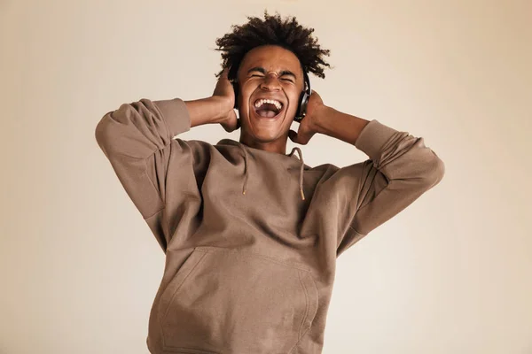 Portrait of a happy young afro american man dressed in hoodie isolated, listening to music with headphones, shouting