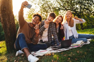 Grup ödev parkta birlikte mutlu multhiethnic öğrenci dizüstü bilgisayar, cep telefonu ile bir selfie alarak kullanan