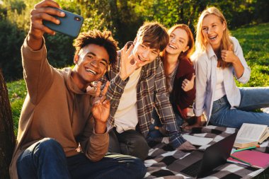 Parkta birlikte ödev olumlu multhiethnic öğrenciler dizüstü bilgisayar, cep telefonu ile bir selfie alarak kullanan Grup