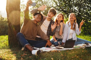 Parkta birlikte ödev neşeli multhiethnic öğrenciler dizüstü bilgisayar, cep telefonu ile bir selfie alarak kullanan Grup