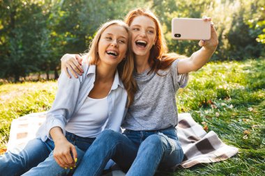 Gülüyor, çimen ve cep telefonu alarak selfie yaz Park'ta dinlenme sırasında otururken rahat kıyafetler giymiş iki neşeli kadın görüntüsünü