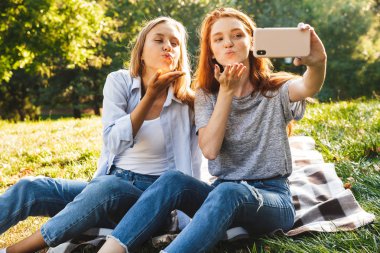 Çimenlerin üzerinde oturan ve hava alarak selfie Smartphone cep telefonu ile yaz Park ederken öp yapma rahat kıyafetler giymiş iki beyaz kız fotoğraf