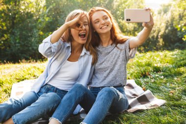 İki güzel kız öğrenciler yaz park ve alma selfie cep telefonuyla çimenlerin üzerinde oturan rahat kıyafetler giymiş görüntüsünü