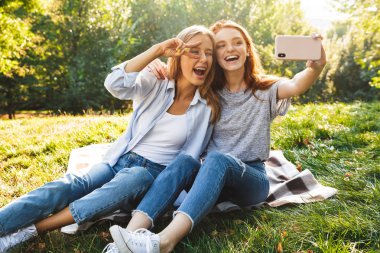 İki güzel kız öğrenciler yaz park ve alma selfie cep telefonuyla çimenlerin üzerinde oturan kot giyen görüntüsünü