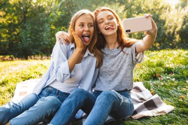 Görüntü yaz Park'ta dinlenme sırasında ot ve alma selfie Smartphone cep telefonu ile oturan kot giyen iki mutlu kadın arkadaş