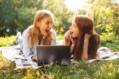 Parkta çim üzerinde yalan ve gümüş dizüstü kullanarak rahat kıyafetler giymiş iki heyecanlı kadın görüntüsünü
