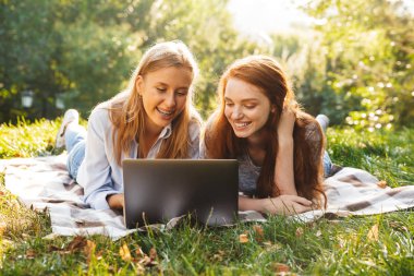 Görüntü parkta çim üzerinde yalan ve gümüş dizüstü kullanarak rahat kıyafetler giymiş iki güzel kadın arkadaş
