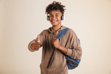 Mutlu bir genç afro Amerikalı adam portresi hoodie içinde başparmak gösteren izole, sırt çantası taşıyan kulaklık ile giyinmiş