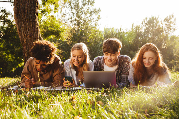 Group of happy multhiethnic students doing homework together at the park, using laptop computer and mobile phone, laying on a grass